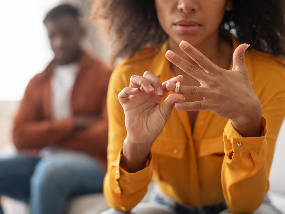 woman takes off her wedding ring, man in background with crossed arms. divorce concept.
